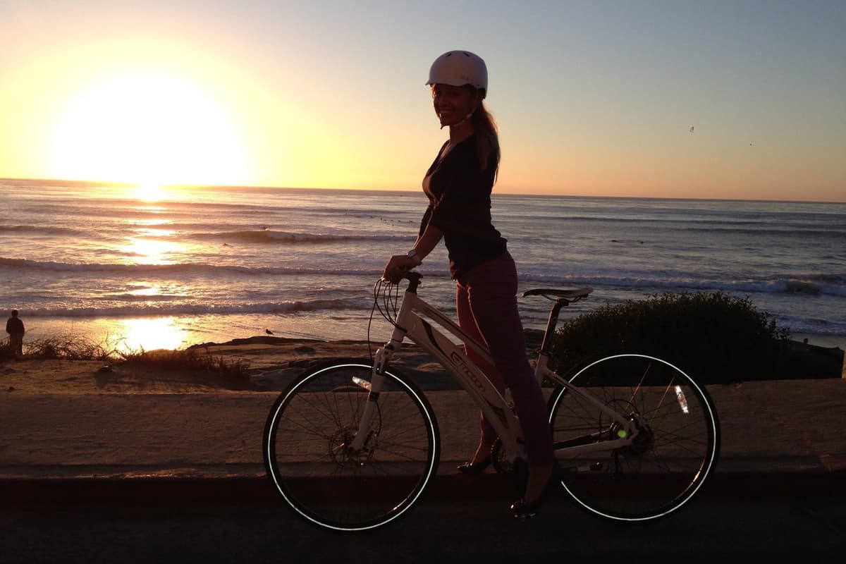 Person standing beside a bicycle at sunset, wearing a helmet and smiling toward the camera, with the ocean and gentle waves glowing in the background.