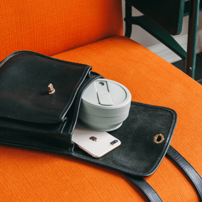 Black leather handbag resting on an orange chair with a reusable travel coffee cup and smartphone inside