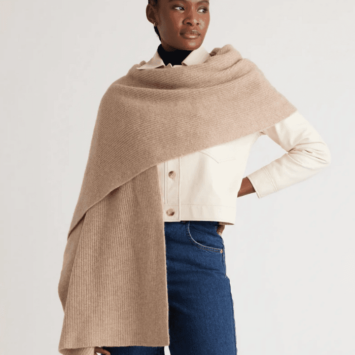 Woman wearing a soft beige knit scarf styled over a cream jacket and blue jeans in a minimalist studio setting