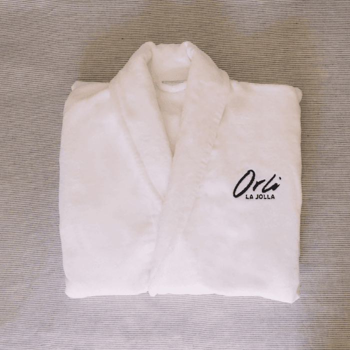 Soft white plush bathrobe embroidered with Orli La Jolla logo, neatly folded on a neutral textured surface