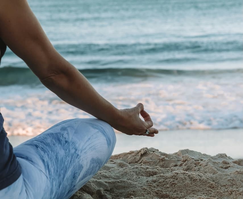 Person seated on a sandy beach in a meditative pose, one arm resting on a bent knee with fingers gently held in a mindfulness gesture, as ocean waves roll in the background.
