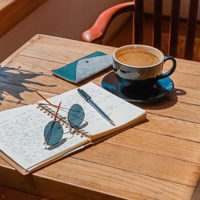 Open notebook with handwritten notes, sunglasses, and pen beside a cup of coffee on a sunlit wooden café table