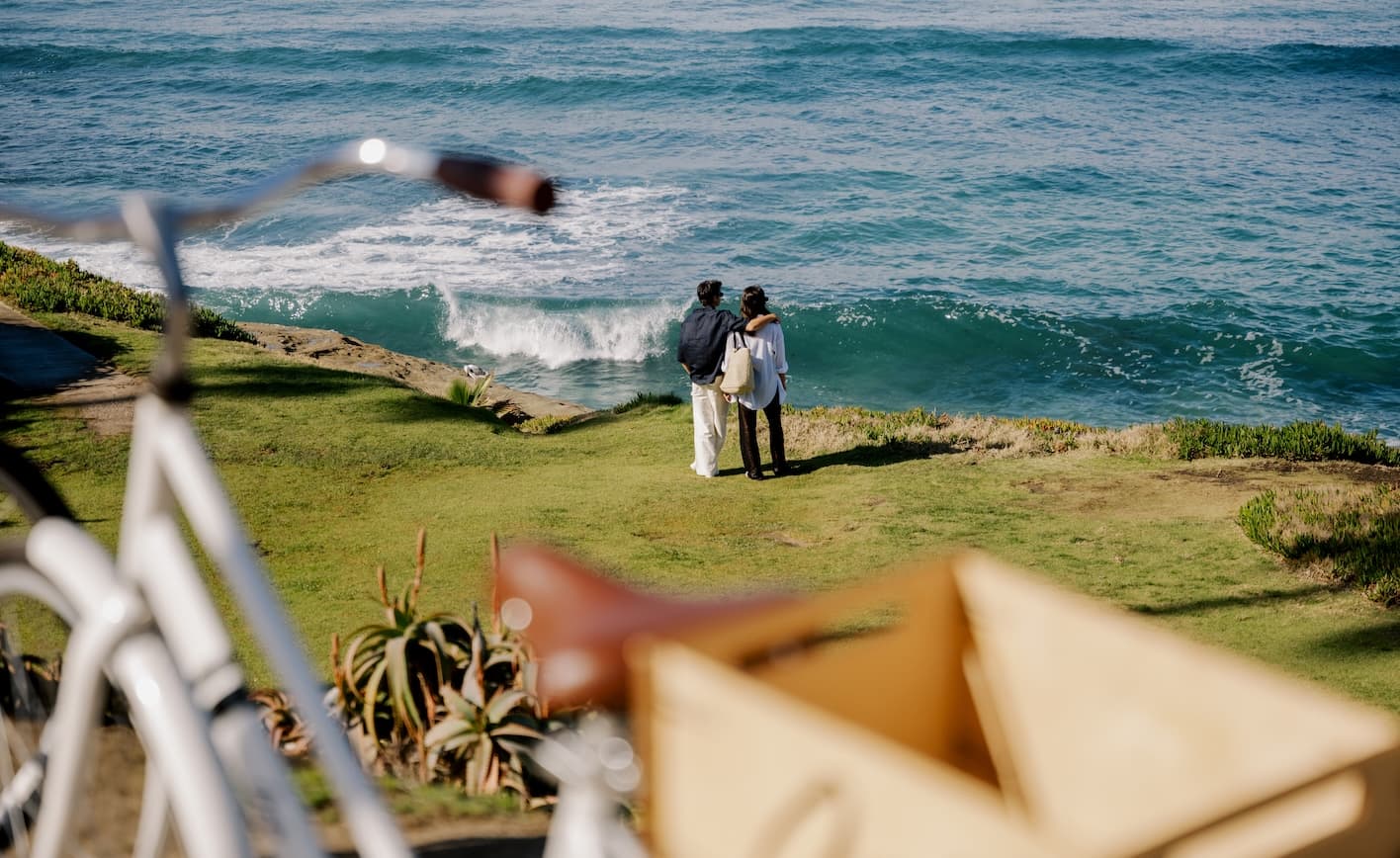 A couple stands arm in arm on a grassy cliff overlooking the ocean as waves roll in below, with soft-focus outdoor furniture in the foreground.