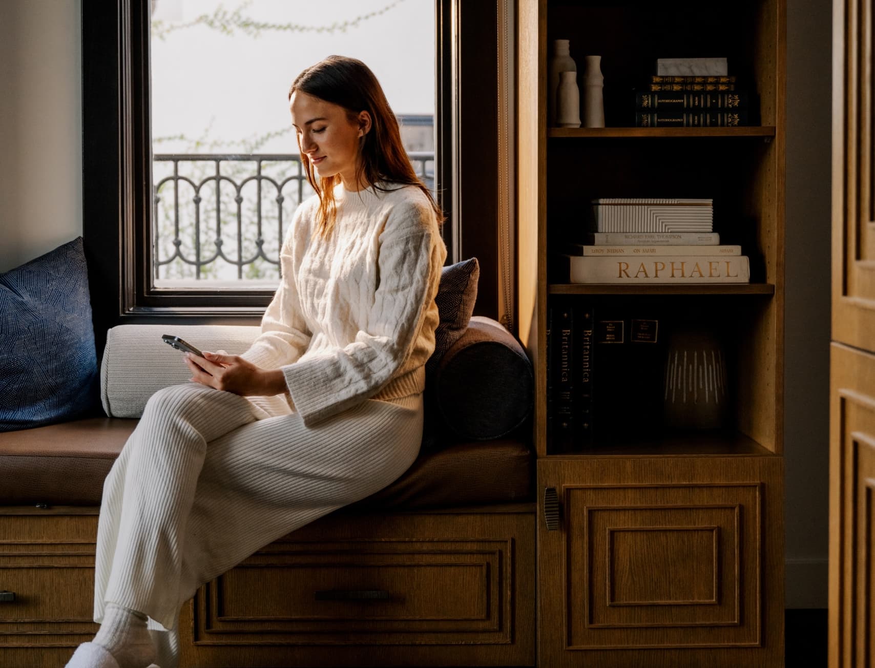 Woman relaxing on a window seat in a luxury hotel room, reading on her phone with natural light and elegant interior design.
