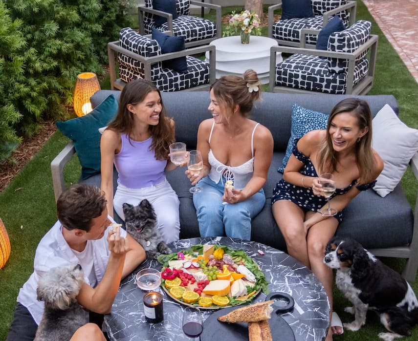 Group of friends relaxing outdoors on a modern patio, sharing wine and a charcuterie board while laughing together, with small dogs nearby, highlighting a casual backyard gathering and social lifestyle.