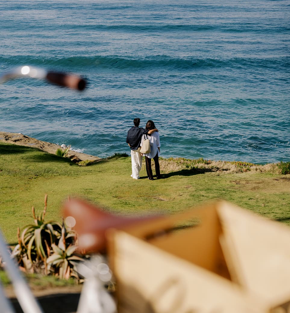 Two people standing close together on a grassy bluff, one with an arm around the other, looking out over rolling ocean waves; the scene is viewed from a distance with softly blurred foreground elements and a calm coastal atmosphere.