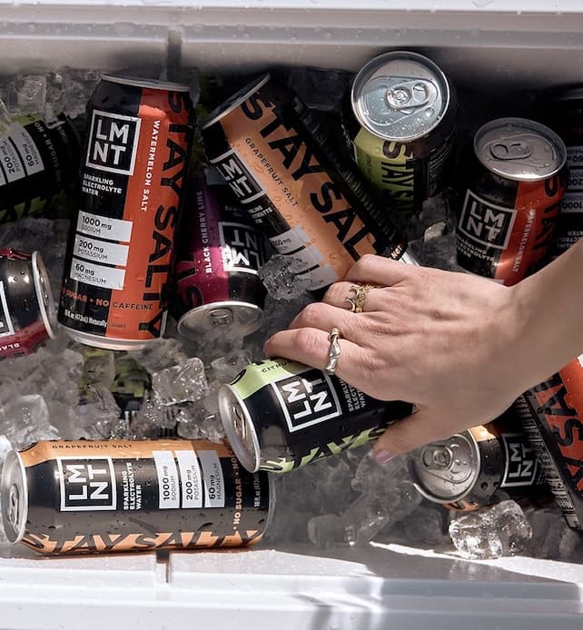 A hand wearing gold rings reaches into a white cooler filled with ice, grasping a black and green can of LMNT sparkling electrolyte water. Several other LMNT cans in different flavors are scattered among the ice, with visible condensation and readable labels, emphasizing a cold, refreshing beverage selection.