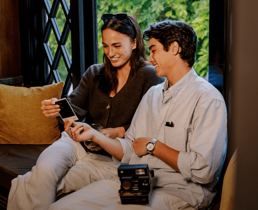Two friends sitting indoors by a window, smiling while looking at a freshly developed instant photo, with a vintage instant camera resting nearby, capturing a warm, candid moment of shared memories and connection.
