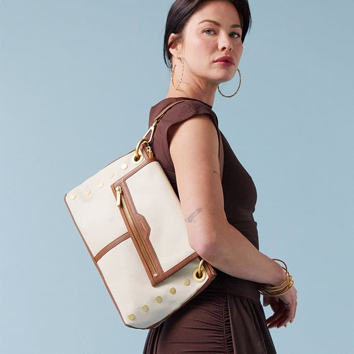 Woman wearing a brown dress carrying a structured cream leather backpack with gold hardware against a minimalist blue background