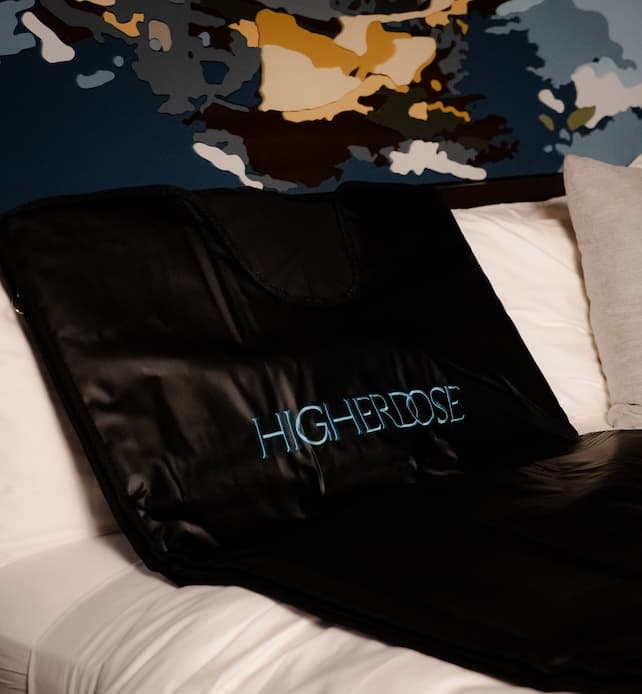 HighERDose infrared sauna blanket folded on a white bed with pillows, showing the embroidered logo in a modern bedroom setting.