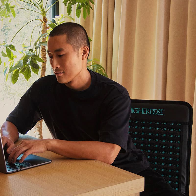 Young man working on a laptop at a wooden desk in a modern, plant-filled interior, seated in an ergonomic office chair with natural light streaming through curtains.