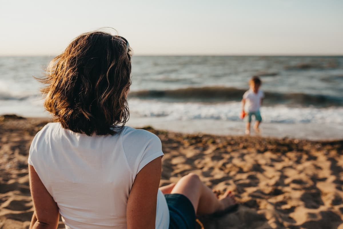 Person sitting on a sandy beach watching a child play near the shoreline, with ocean waves in the background during golden-hour light.