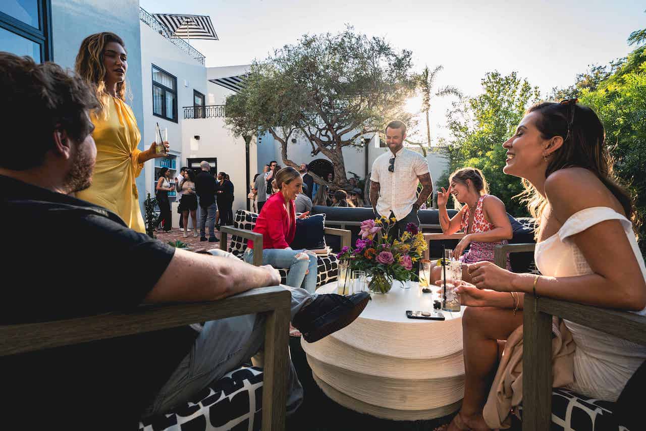 Guests socializing at an outdoor courtyard gathering at Orli La Jolla, seated around a table with cocktails in warm evening light.