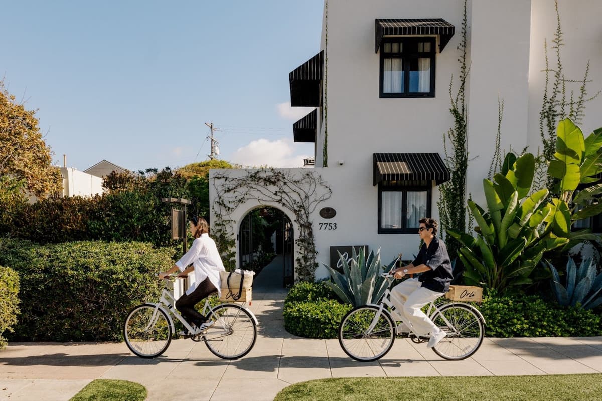 Two people riding bicycles past a white, ivy-covered building entrance on a sunny residential street.