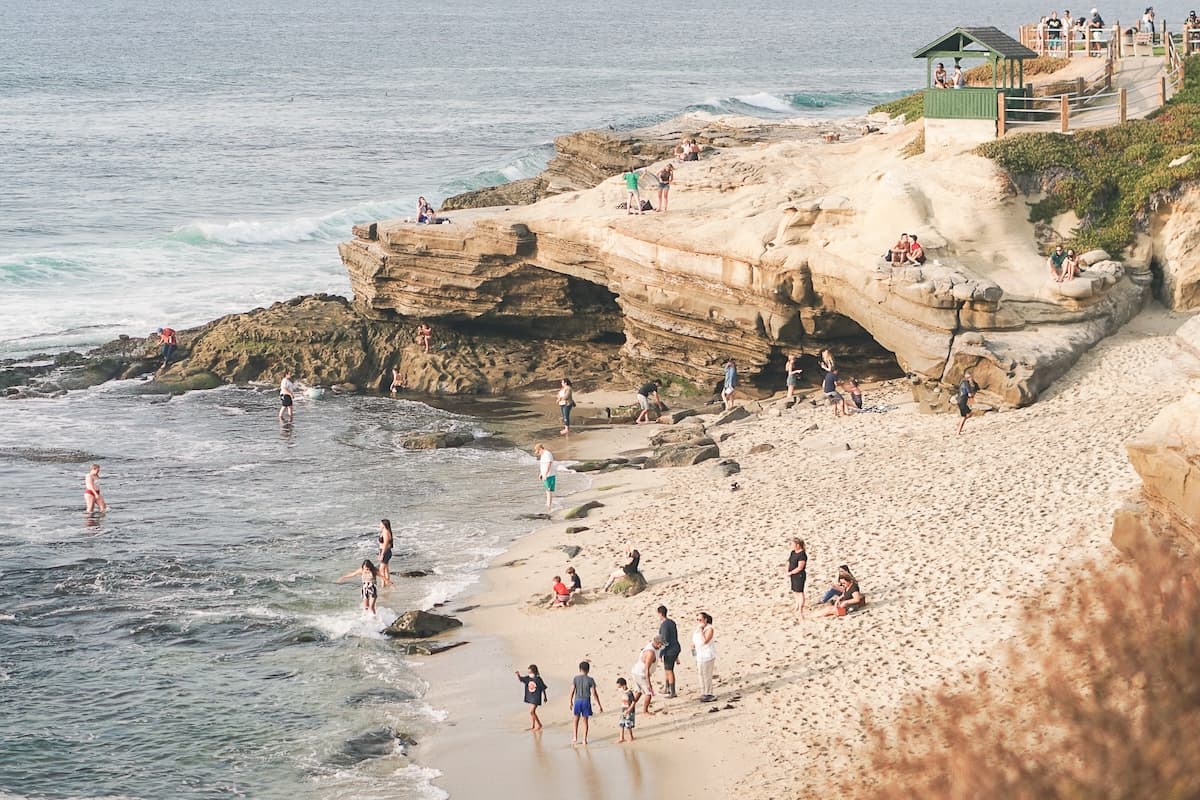 Sandy beach bordered by layered coastal cliffs and rocky shoreline, with people walking, relaxing, and exploring near the water’s edge.