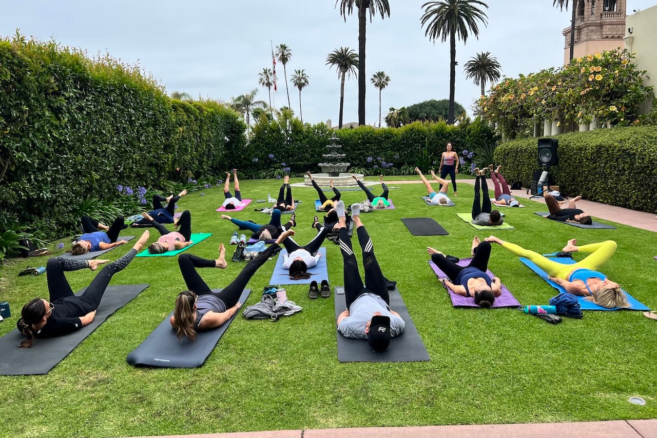 A group participates in an outdoor fitness class on yoga mats spread across a manicured lawn, following an instructor near a fountain. Tall palm trees, hedges, and flowering plants surround the garden.